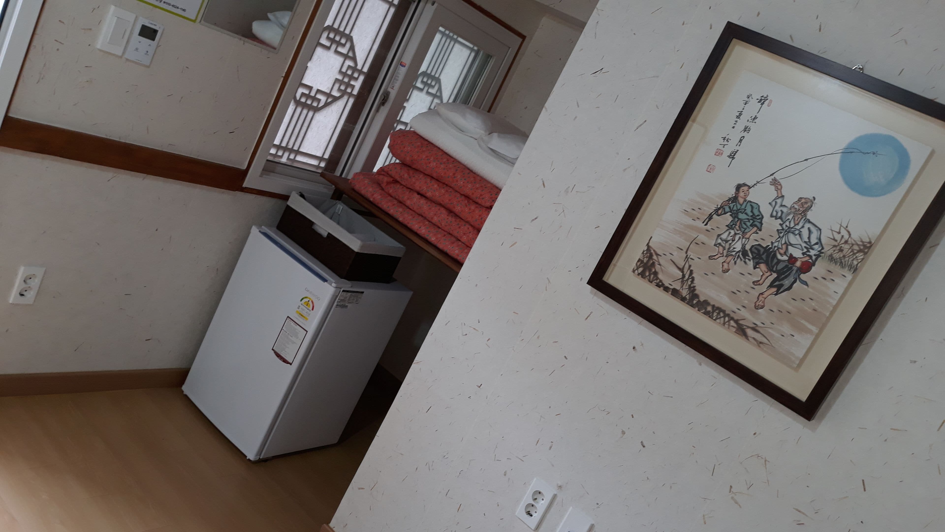 A corner of a room with a white refrigerator, a small heater, and a framed artwork on the wall.