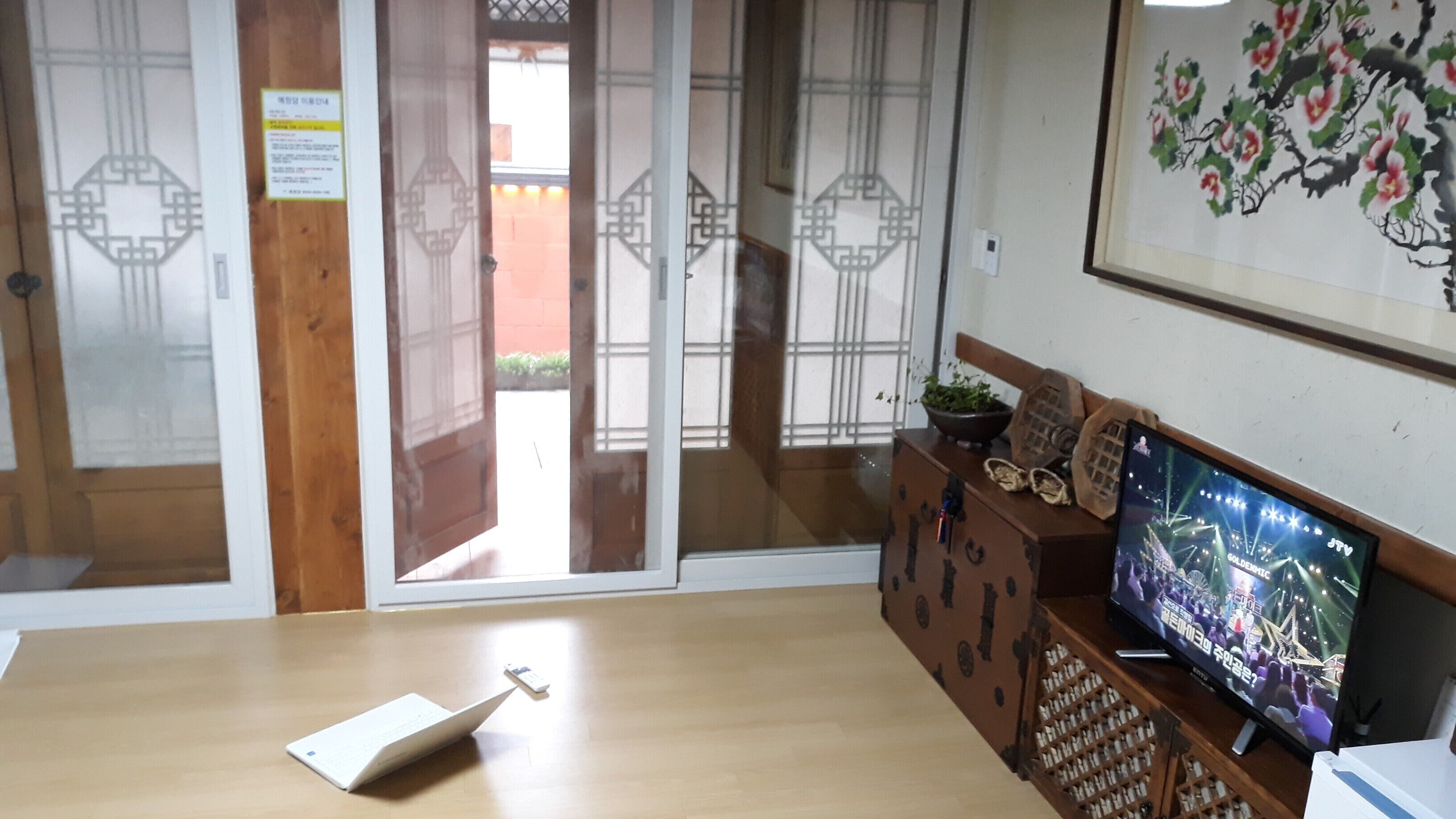 A room with a wooden table, a television displaying a show, a sliding door with a traditional design, and a wall with a floral painting.