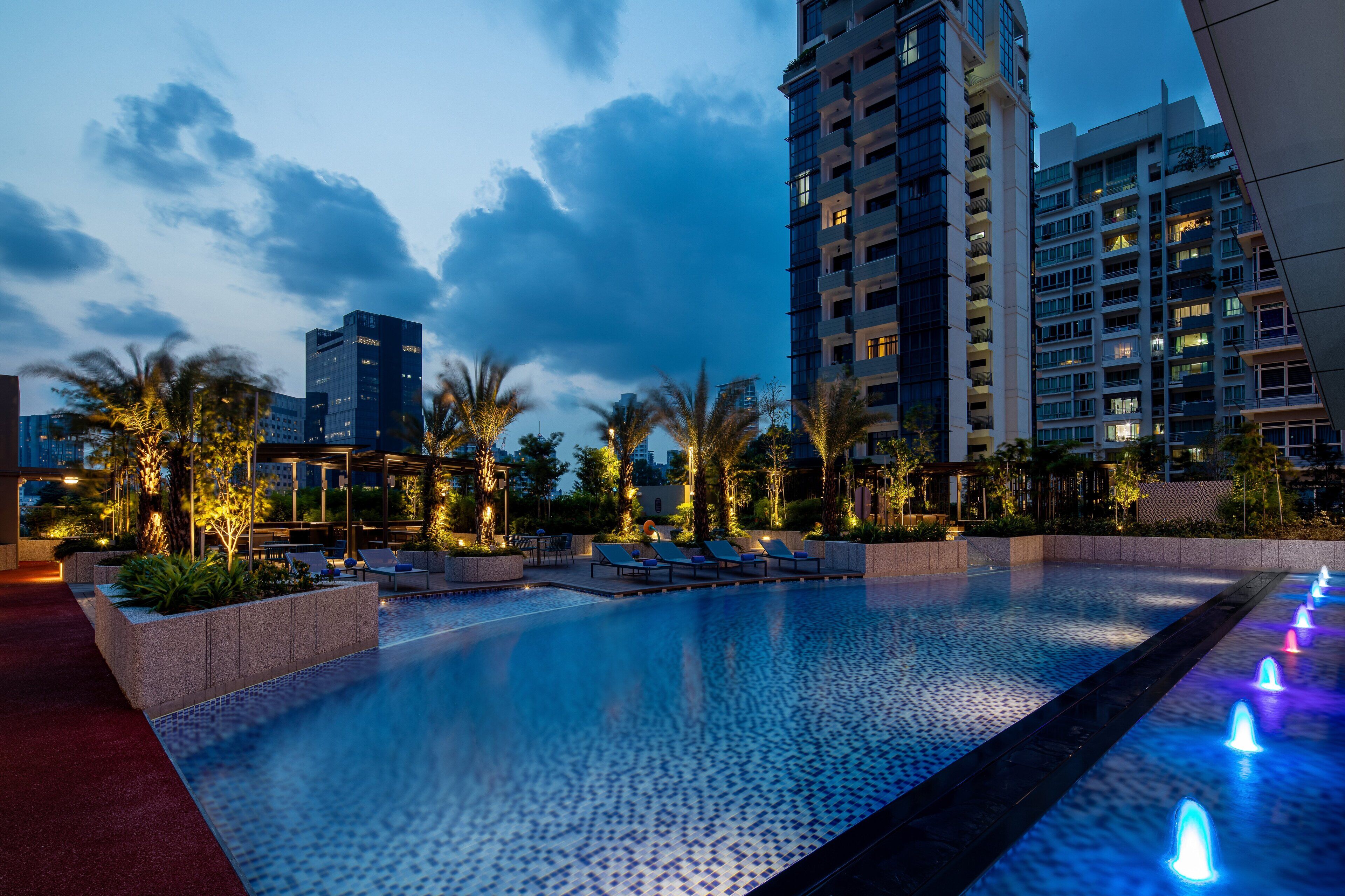 A swimming pool with water features, surrounded by palm trees and modern buildings.