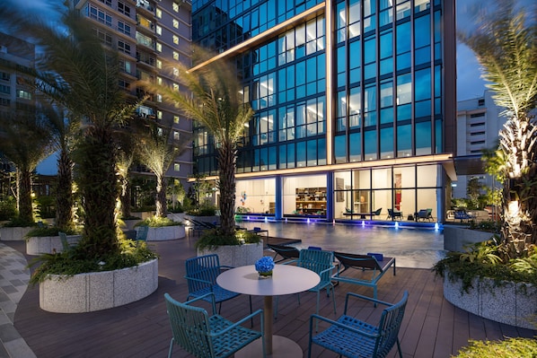 Un patio moderno de hotel con mobiliario exterior, palmeras y una zona de piscina.