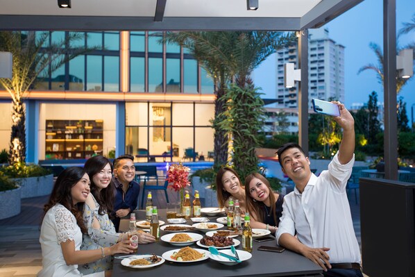 Un groupe de personnes est assis à une table en plein air dans un restaurant, d’où l’on peut admirer un bâtiment moderne et des palmiers.