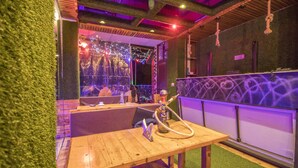 A wooden table with a hookah, a bar area with graffiti, and a stage with lights in the background. - The Siddhartha (Dharamshala)