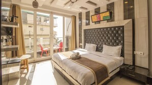 A hotel room with a large bed, a ceiling fan, a TV mounted on the wall, and a balcony with a red chair. - The Siddhartha (Dharamshala)