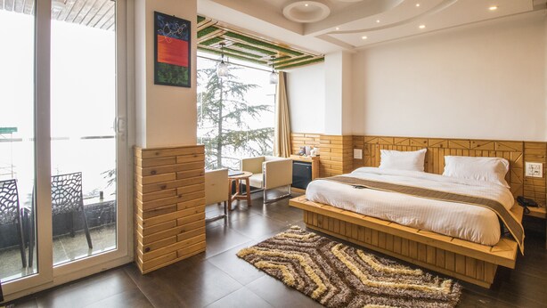 A modern bedroom with a wooden headboard, a large bed, a seating area with a chair and table, and a view through a glass door. - The Siddhartha (Dharamshala)