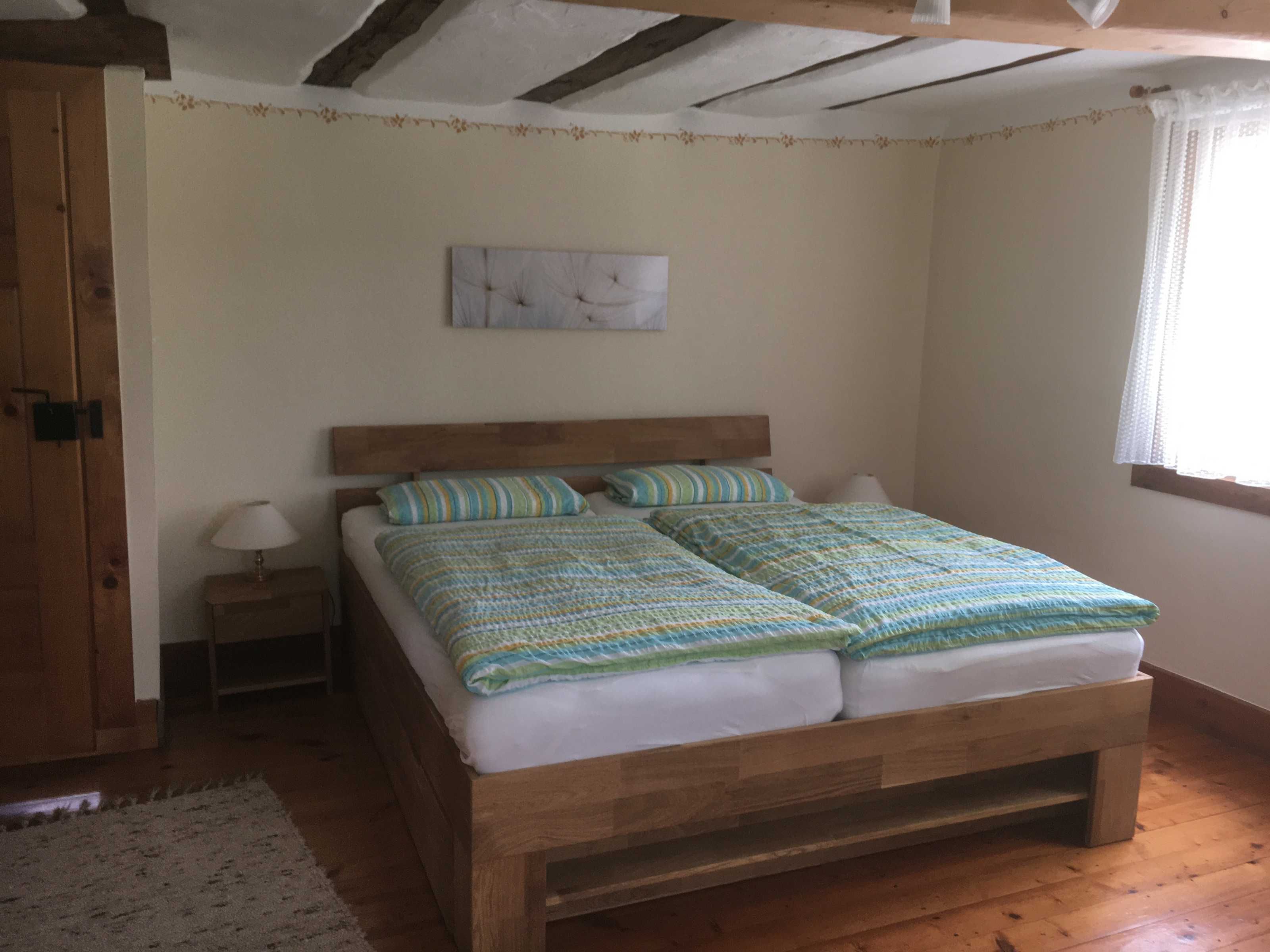 Un lit en bois avec une tête de lit, deux oreillers et une couverture, dans une pièce dont le sol est recouvert de planches de bois et dont un des murs est blanc.