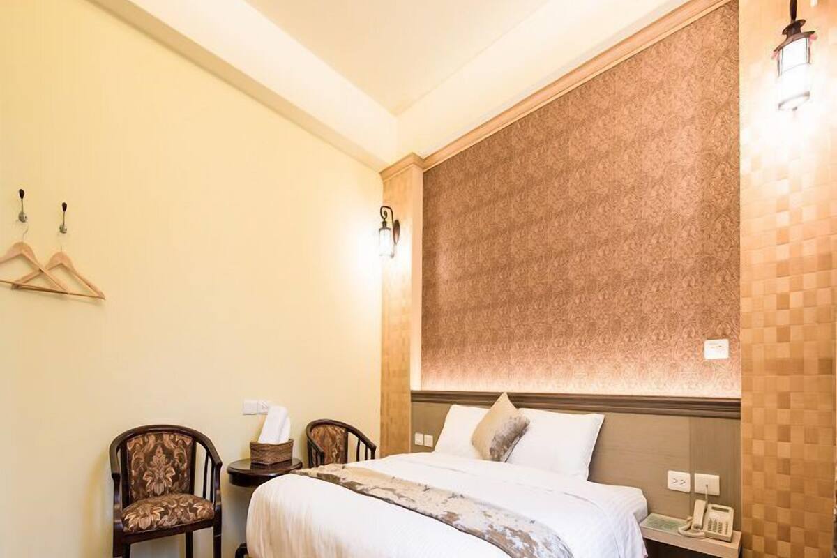 A hotel room with a large bed, a bedside table with a lamp, a chair, and a wall-mounted light fixture.