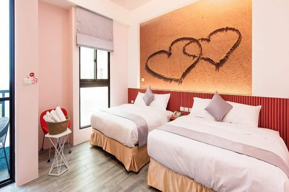 A hotel room with two beds, a red chair, a small table with a basket, and a heart-shaped artwork on the wall.