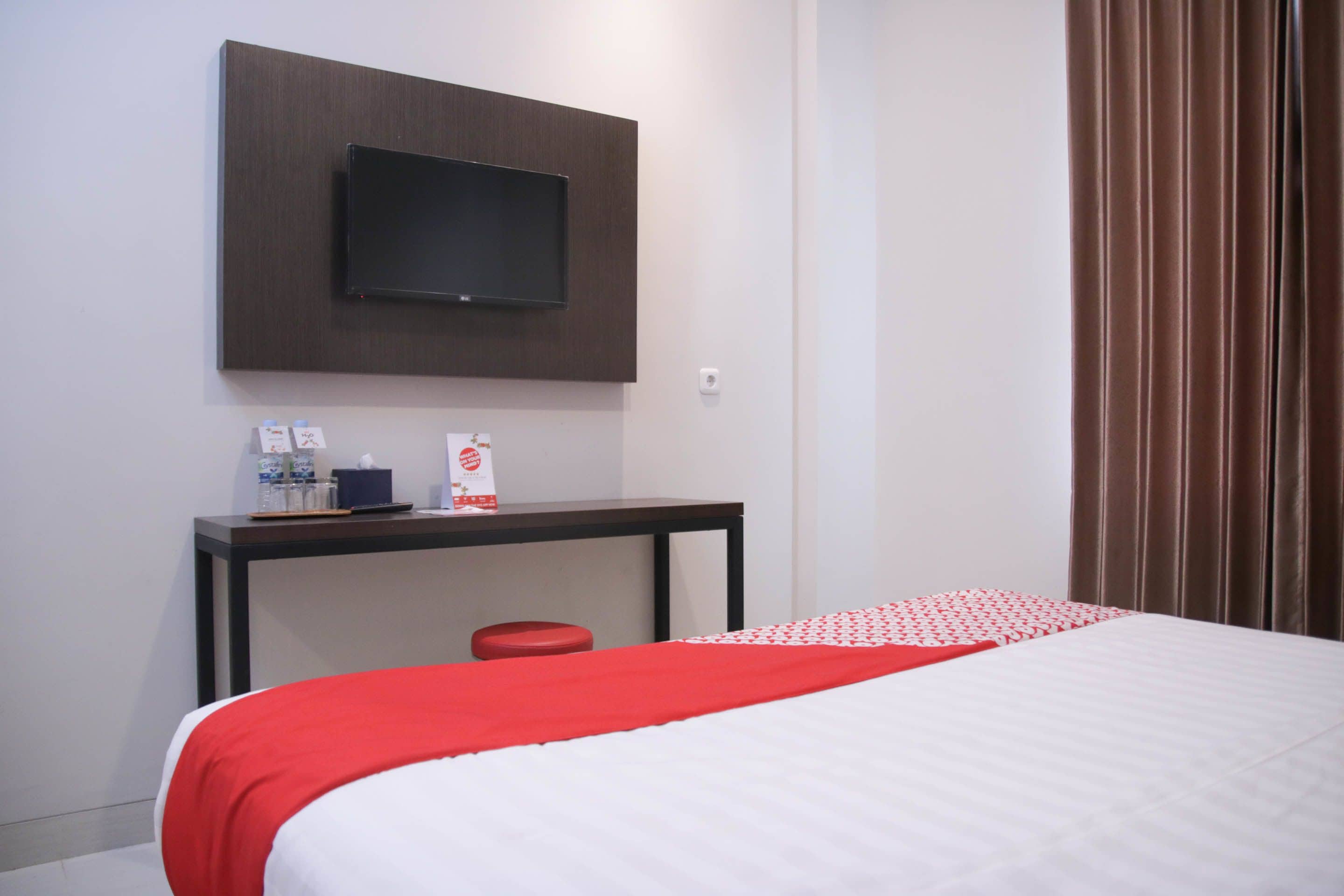 A hotel room with a bed, a TV mounted on the wall, a small table with a red stool, and a window with curtains.