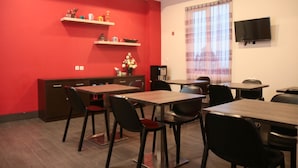 Un espace repas avec des murs rouges, plusieurs tables et chaises, un téléviseur à écran plat fixé au mur et une fenêtre avec des rideaux.