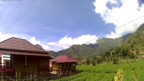 A rural setting with two buildings with red roofs, surrounded by lush green fields and mountains in the background. - Bale Sembahulun Cottages & Tent (Sembalunbumbung)