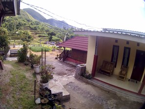 A house with a red roof, a porch with chairs, and a view of a green valley with mountains in the background. - Bale Sembahulun Cottages & Tent (Sembalunbumbung)