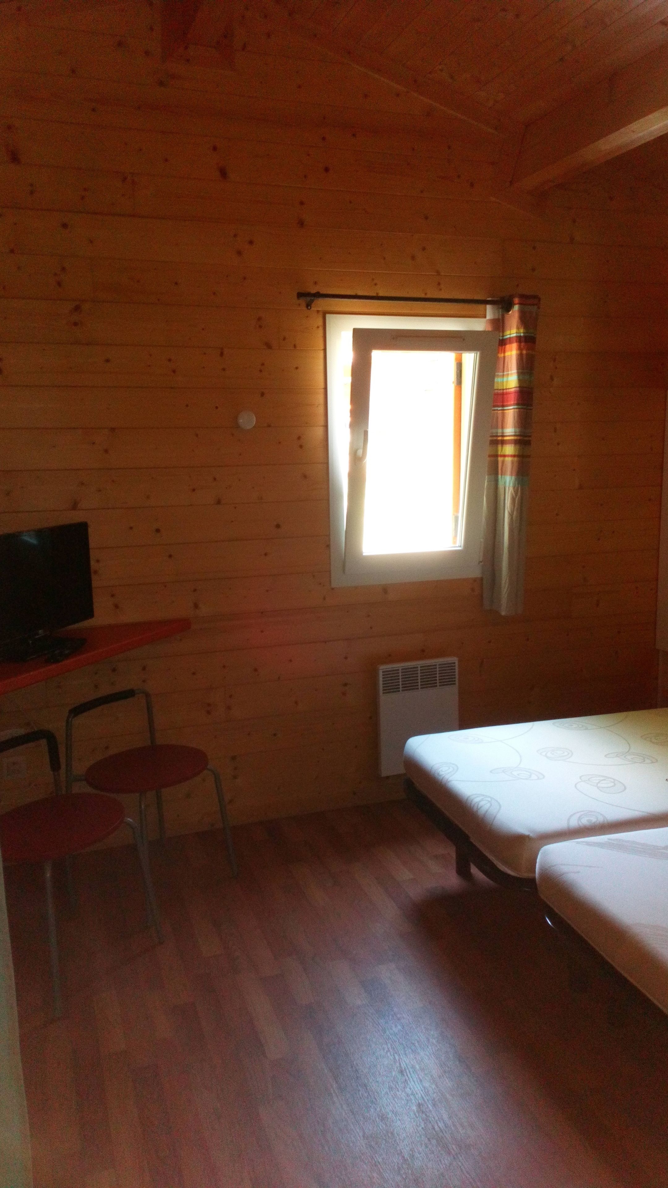 A small, cozy room with wooden walls and a sloped ceiling. It includes a bed, a desk with a chair, and a television.