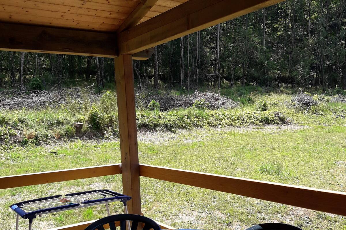 A covered outdoor seating area with a table and chairs, a metal drying rack, and a view of a grassy field and trees.