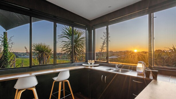 Una cocina moderna con ventanas amplias, dos taburetes de bar y vistas al atardecer.