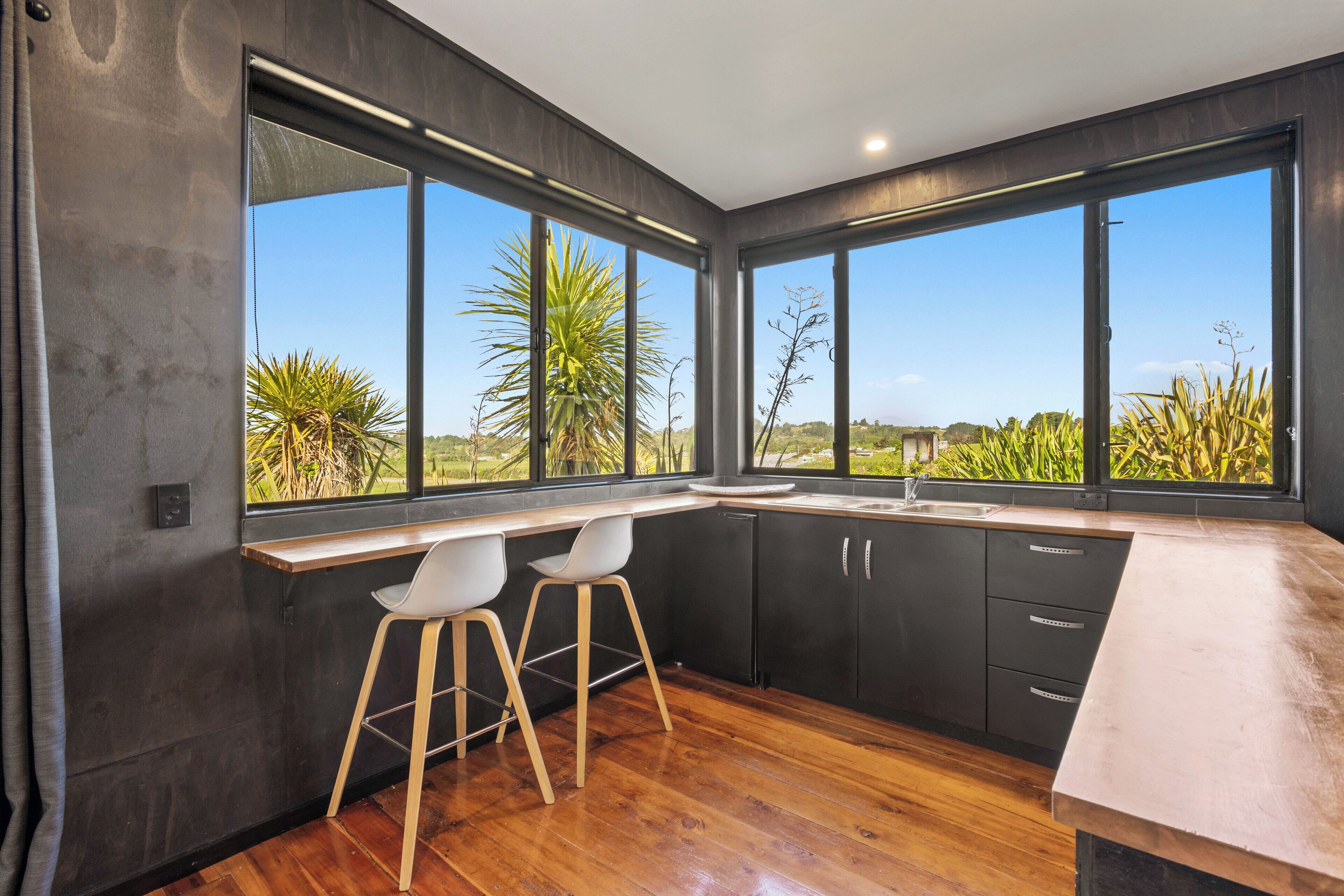 A modern kitchen with large windows, wooden flooring, and two white bar stools.