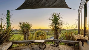 Terraza de madera con dos sillas y una mesa, con vista a un paisaje verde con palmeras y cielo despejado.