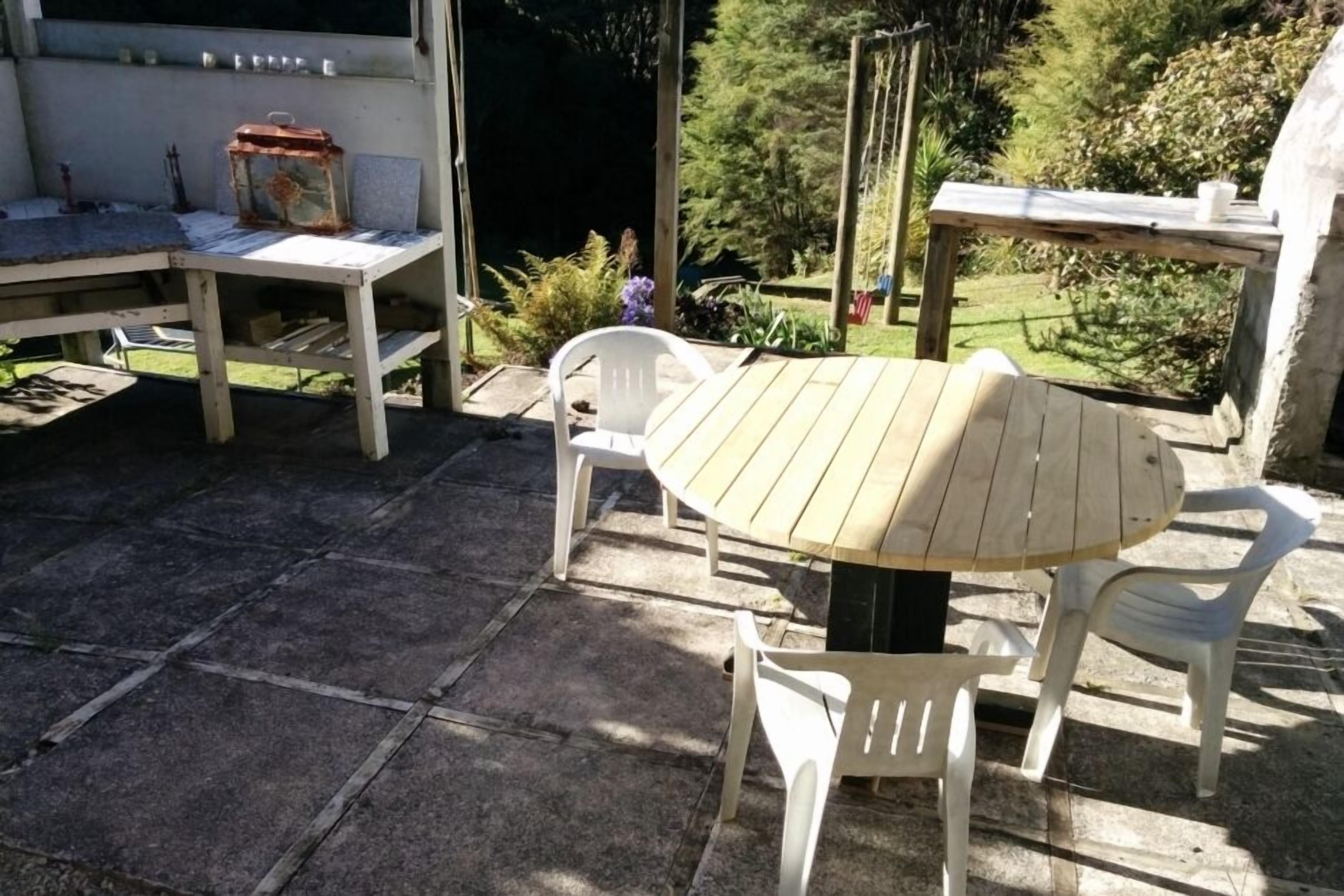 An outdoor patio with a round wooden table and white plastic chairs, surrounded by stone tiles.