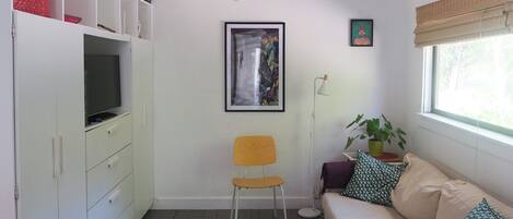 Una sala de estar con un sofá, una silla, una mesa de centro con un libro, un televisor sobre un mueble, un cuadro en la pared y una ventana con una planta.