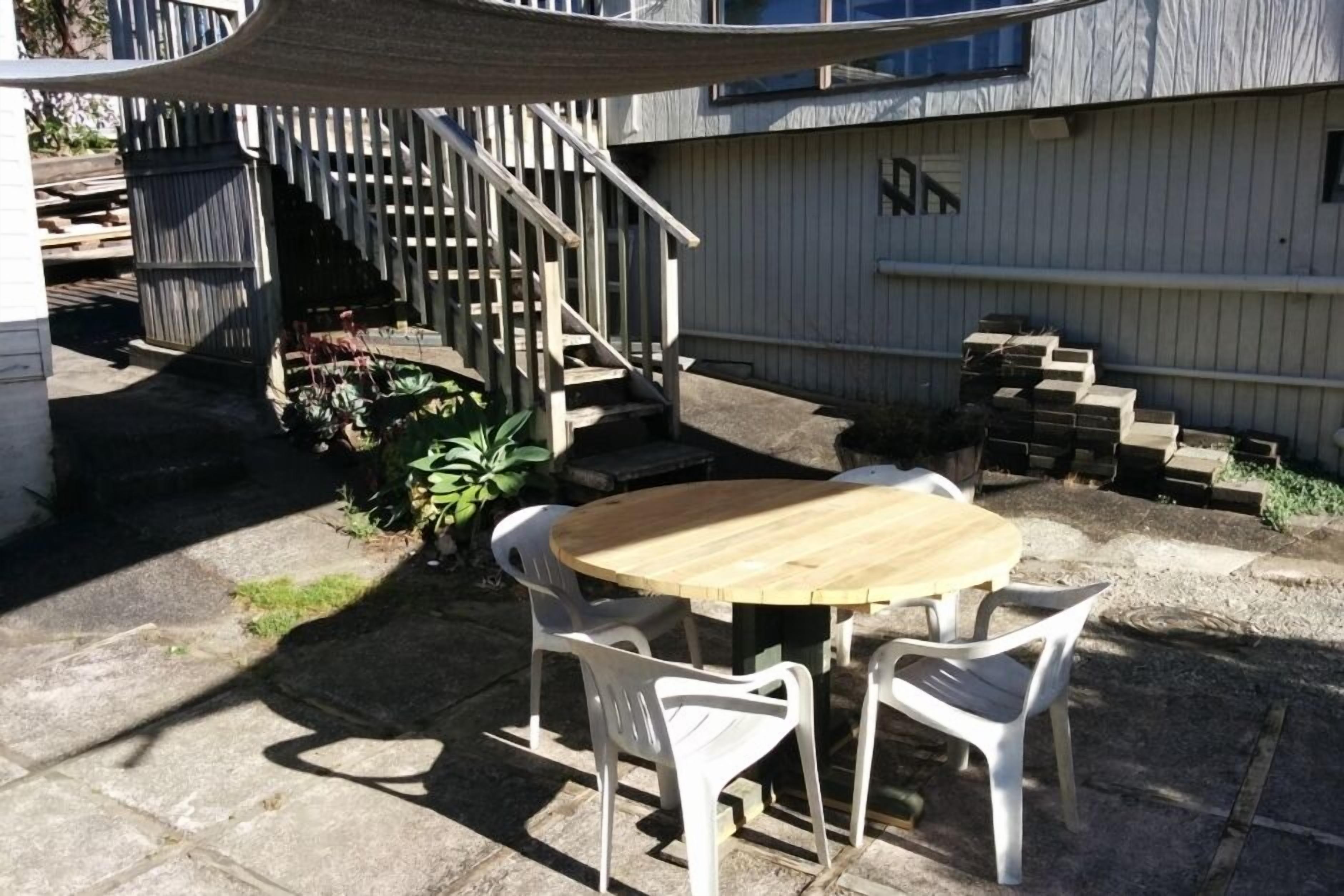 Un patio con una mesa redonda de madera y sillas de plástico blanco, una escalera de madera y un edificio blanco con una ventana y una puerta.