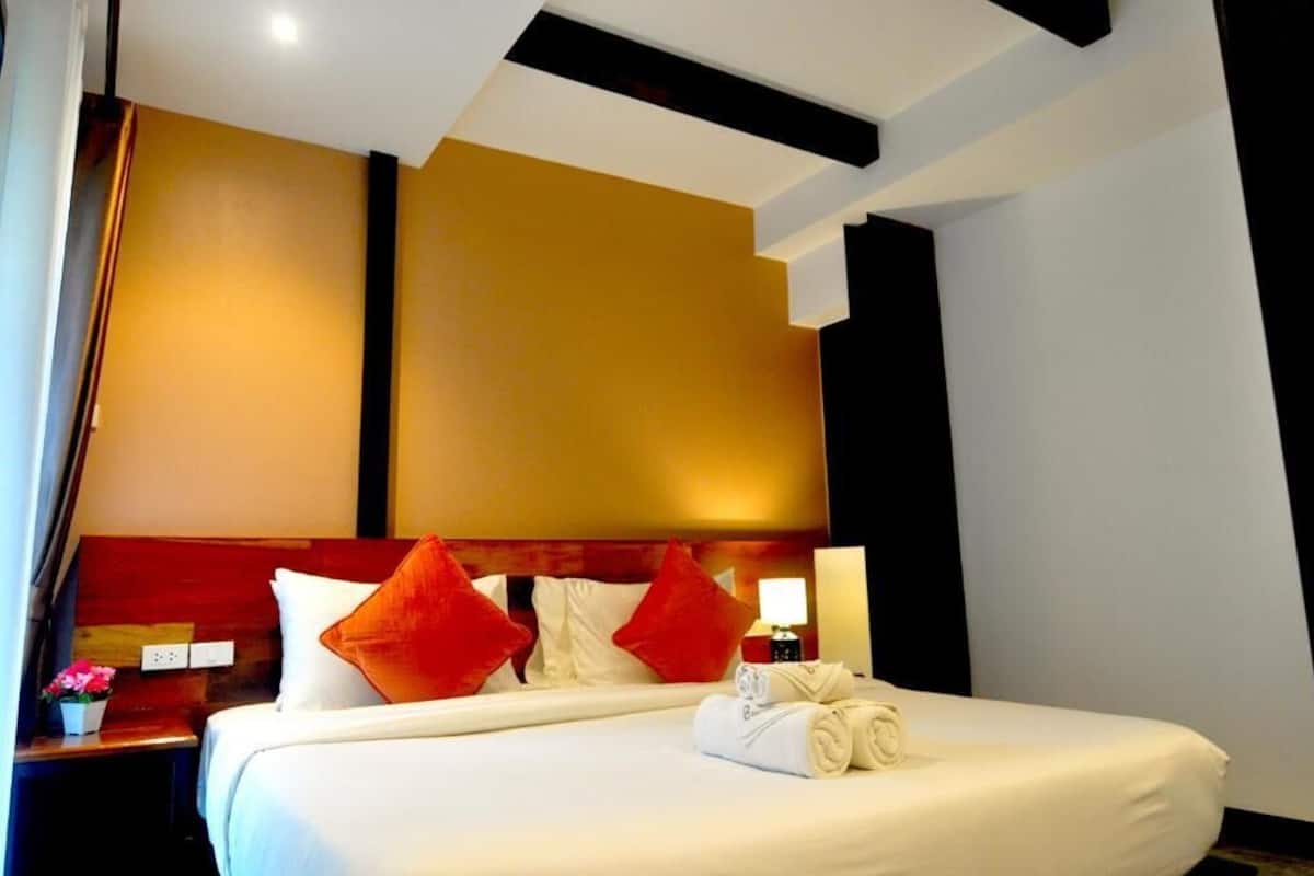 A hotel room with a large bed, two red pillows, a bedside table with a lamp, and a neatly made bed with a rolled towel.