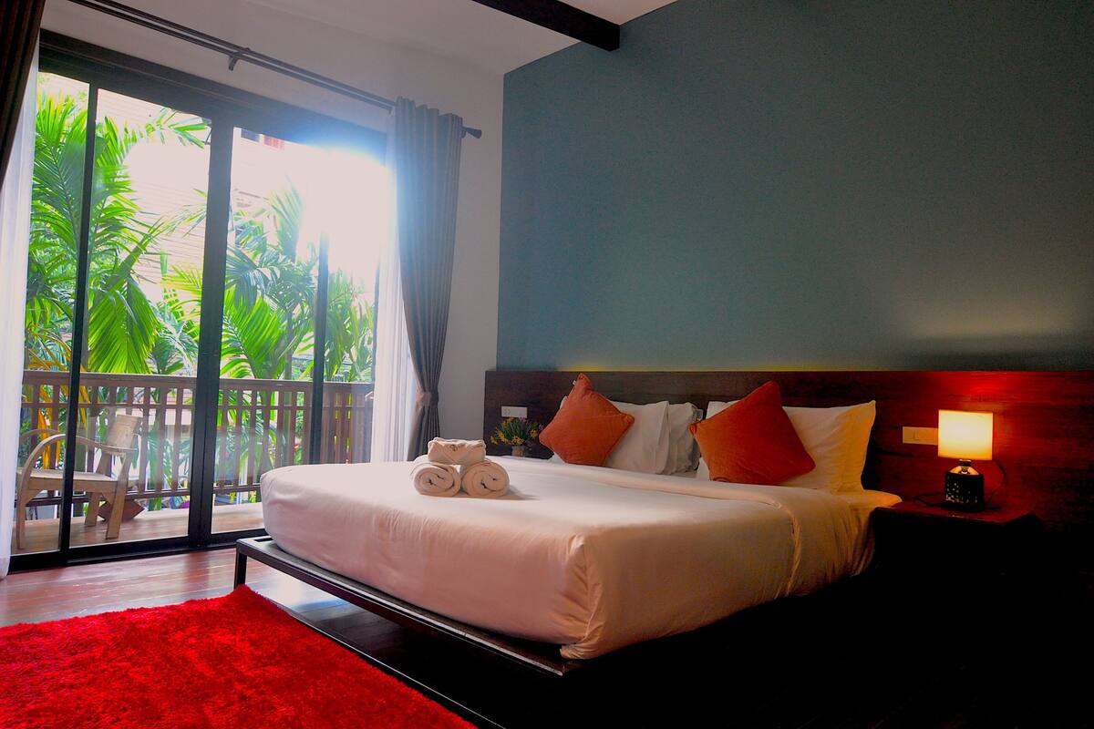 A hotel room with a large bed, a red rug, a nightstand with a lamp, and a balcony with a view of palm trees.