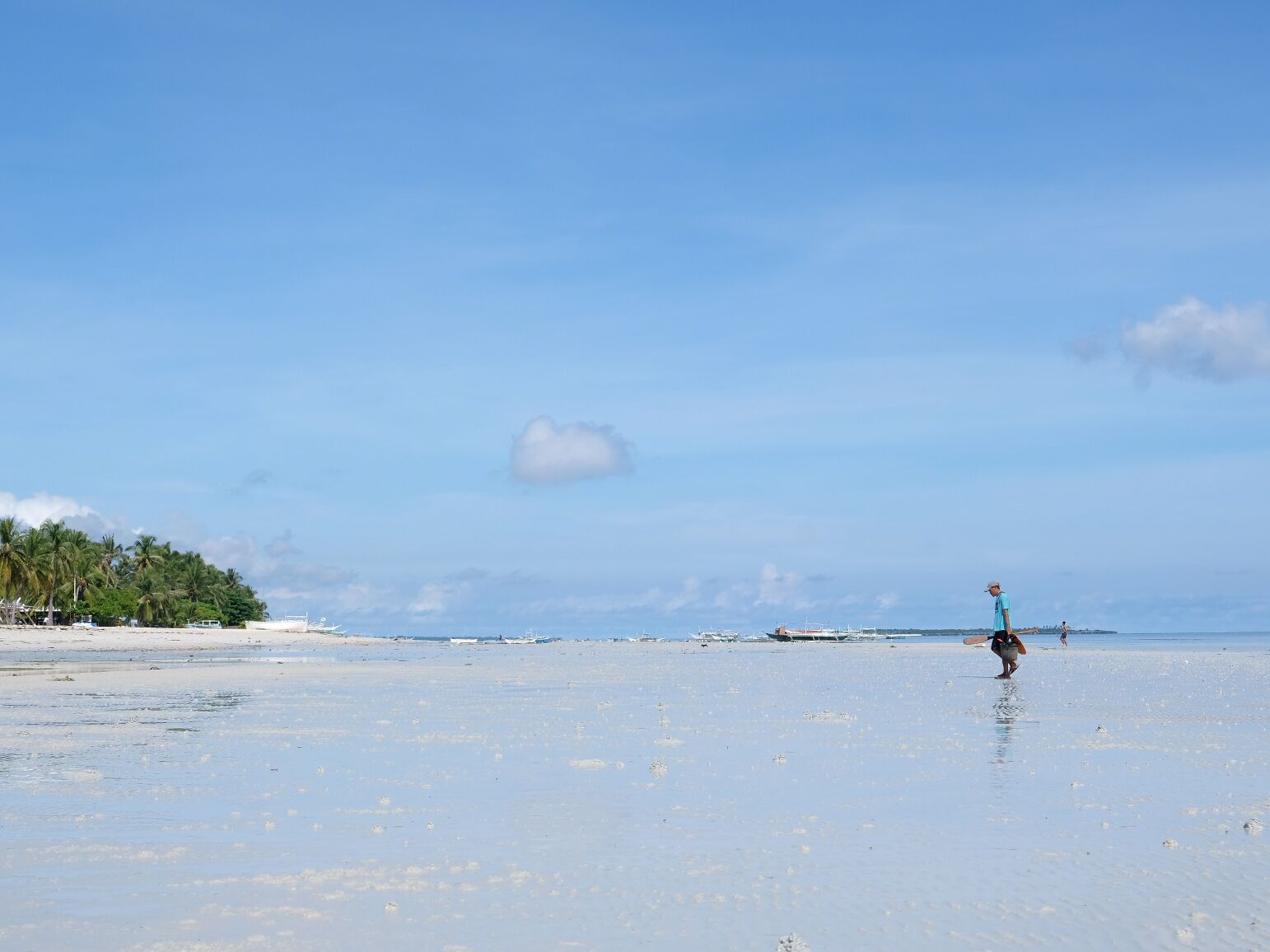 Una playa con una persona caminando, palmeras y barcos a lo lejos.