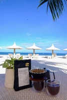 Una mesa con un menú, una cafetera y una taza de café en una playa con sombrillas y sillas de descanso.