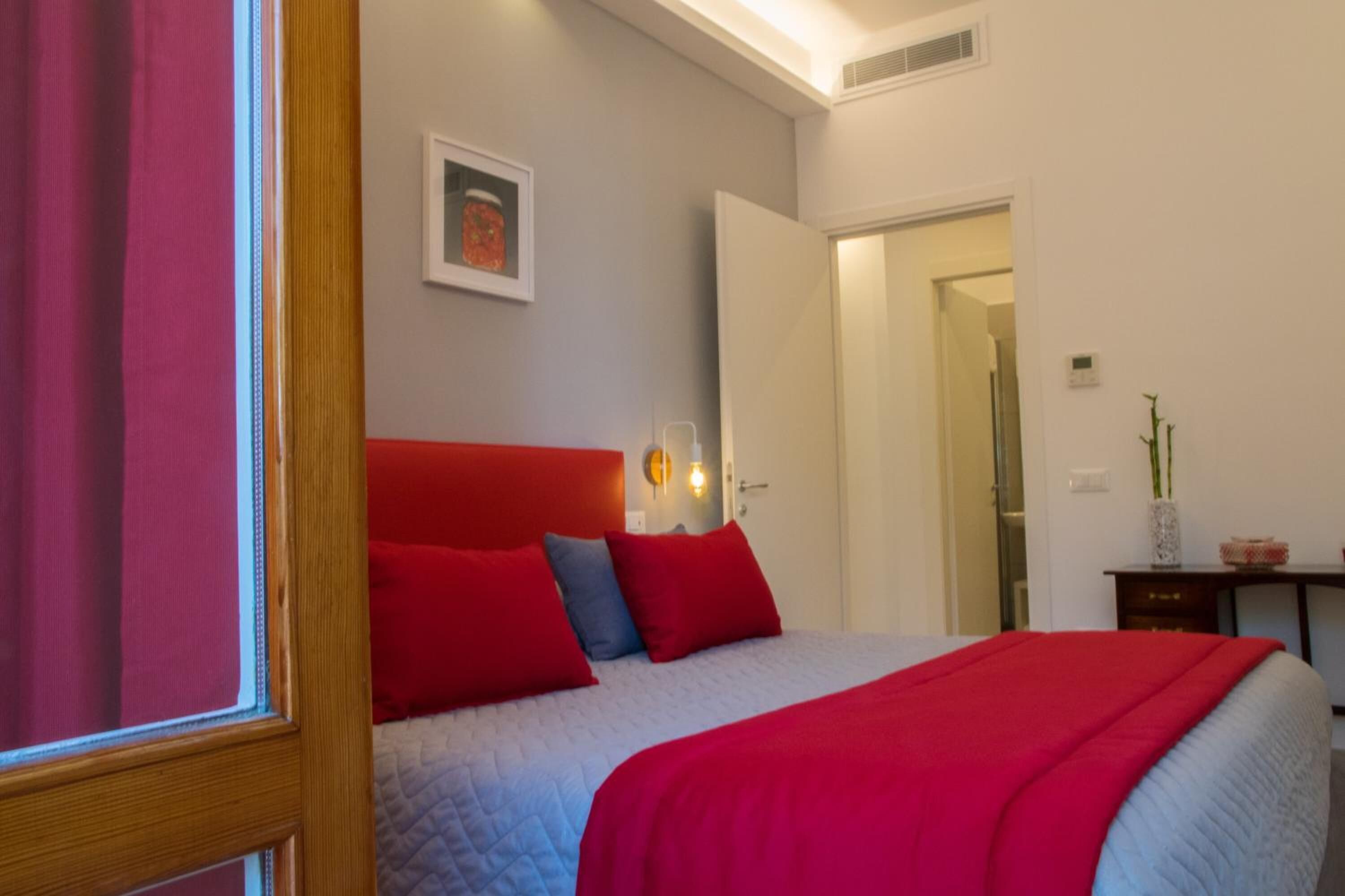 A bedroom with a bed, red and blue pillows, a red headboard, a bedside table with a lamp, and a framed picture on the wall.