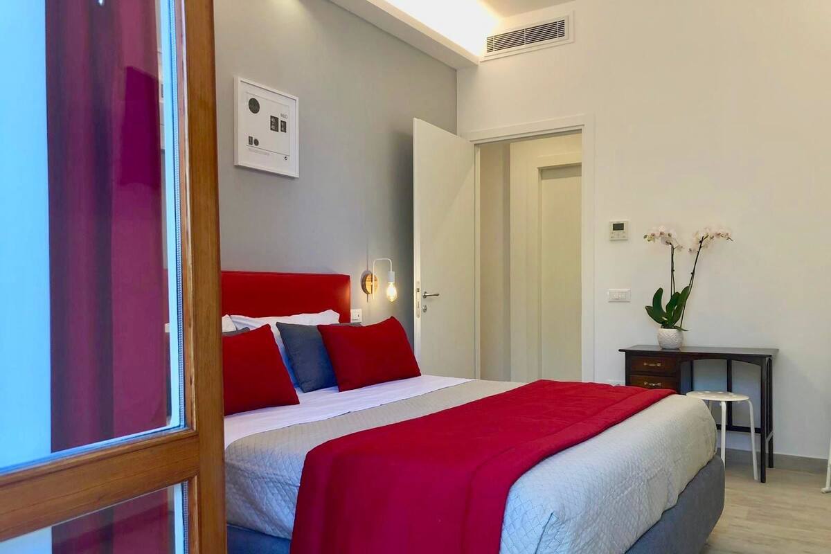 A hotel room with a bed, a bedside table, a desk, a chair, and a potted plant.