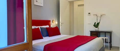 A hotel room with a bed, a bedside table, a desk, a chair, and a potted plant.