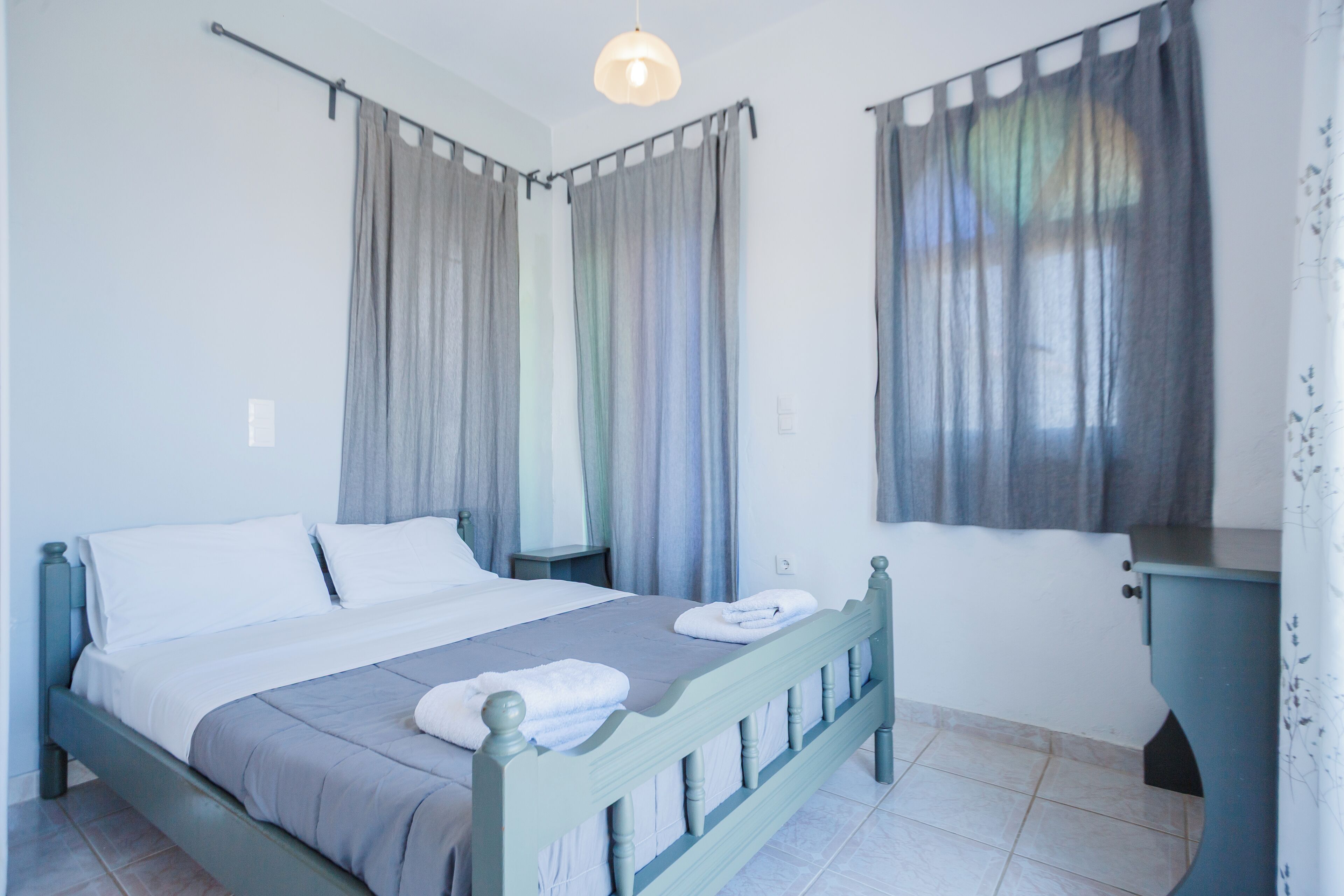 A neatly made bed with white linens and a grey blanket, a bedside table with a lamp, and a window with curtains.