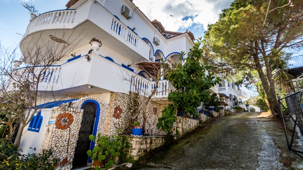 Ein weißes Gebäude mit blauen Akzenten und einer Steinfassade, mit einem geschwungenen Balkon und einer schmalen Straße, die darauf zuläuft.