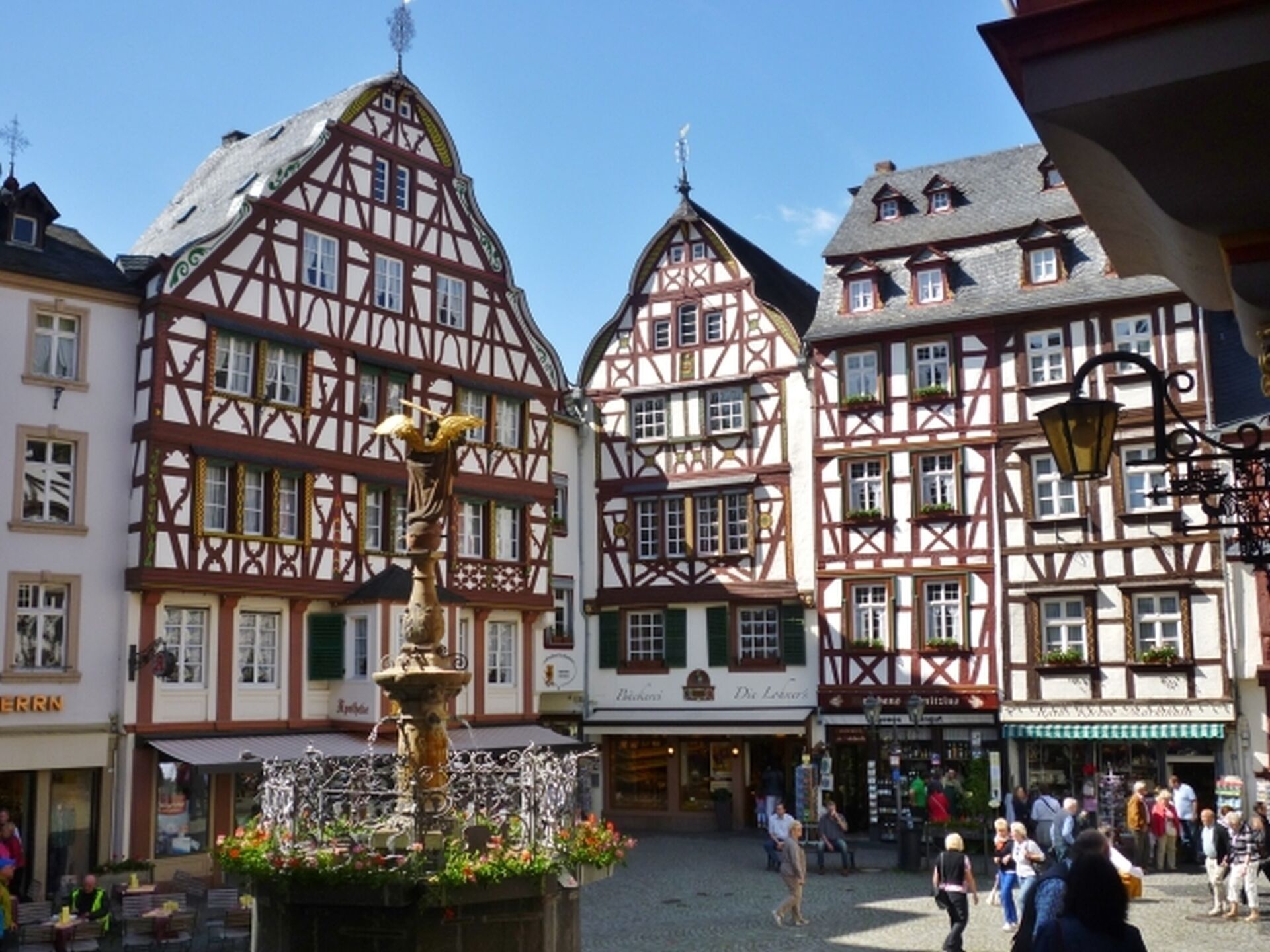 A traditional European town square with half-timbered buildings, a central fountain, and people walking around.
