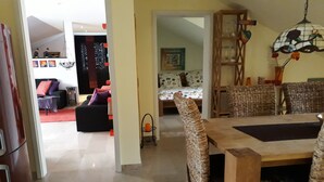 Ein Essbereich mit Korbstühlen und einem Holztisch, ein Wohnzimmer mit einer Couch und einem roten Teppich sowie eine Küchenzeile mit einem Kühlschrank.