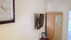 A room with a wall-mounted clock, a wooden cabinet with a mirror, and a small table with a kettle. - Standard Way Lodge (Livingstone)