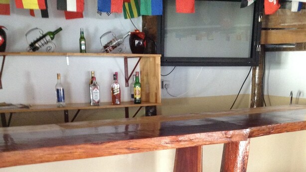 A bar area with a wooden counter and four copper stools. - Standard Way Lodge (Livingstone)