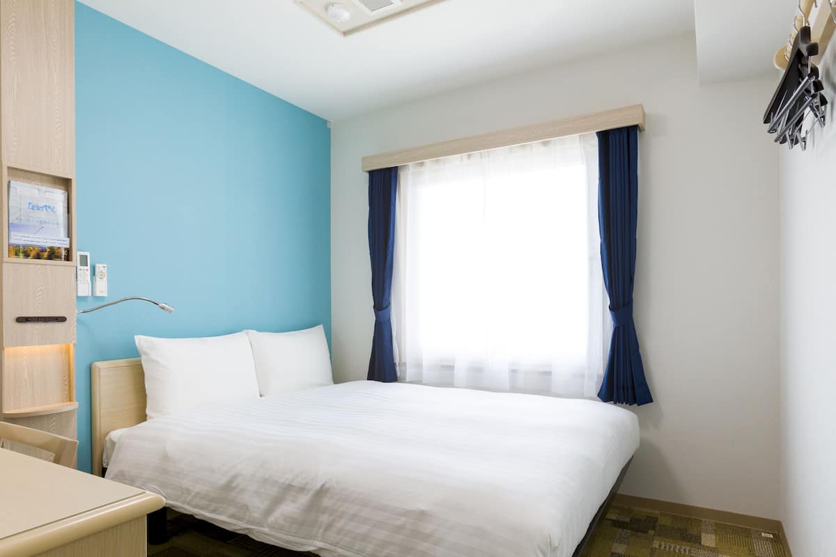 A hotel room with a bed, a desk, a nightstand, a window with blue curtains, and a wall-mounted clock.
