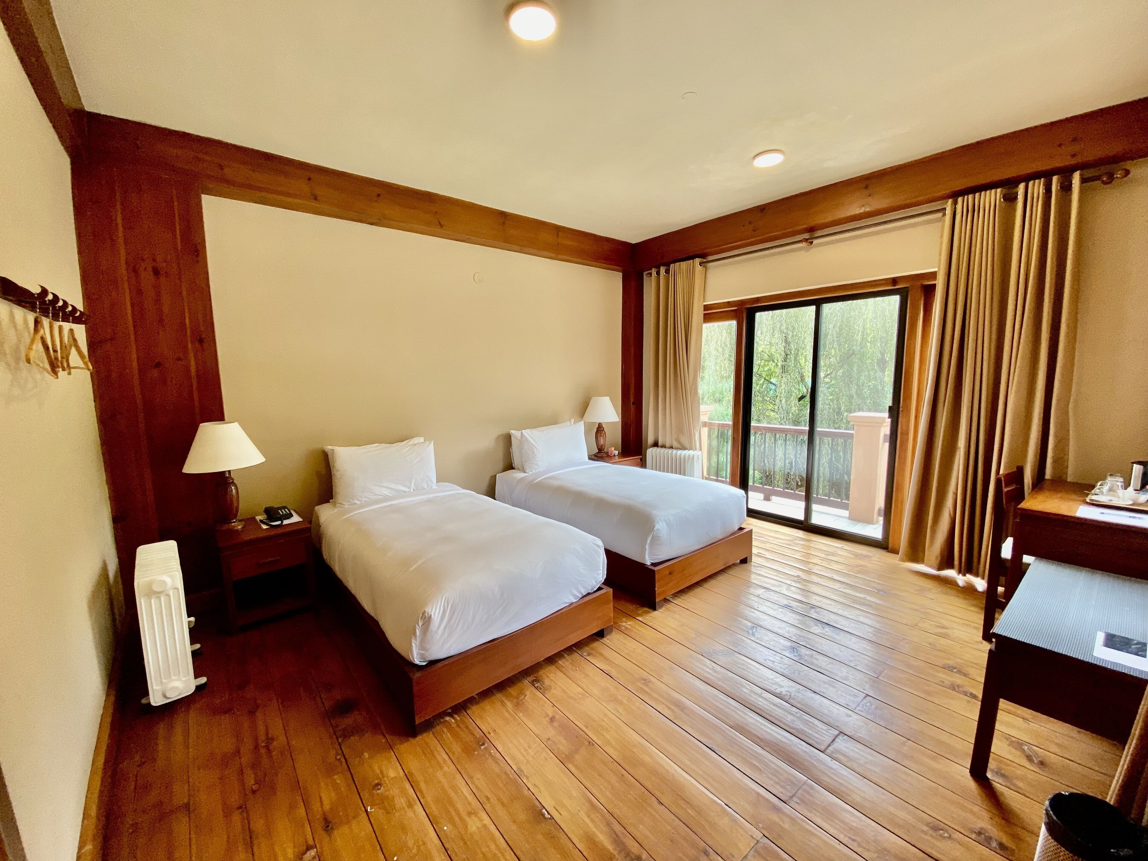 A hotel room with two beds, wooden flooring, a desk, and a view of the outdoors.