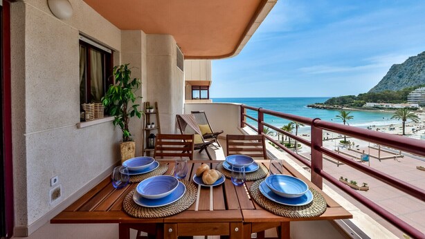 Ein Balkon mit einem Holztisch, gedeckt für das Essen, mit Blick auf einen Strand und eine gebirgige Landschaft.