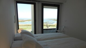 A bedroom with two beds, a chair, and a view of the beach and ocean through large windows. - A beautiful one bedroom apartment - right on the waterfront in the Ria Formosa. (Fuseta)
