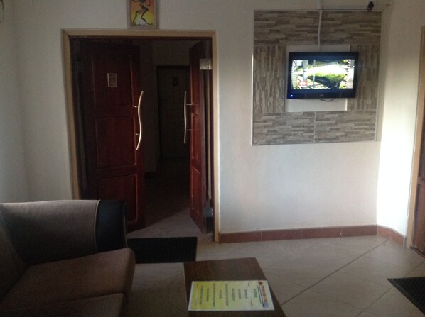 A living room with a sofa, a coffee table with a document, a TV mounted on a stone wall, and a door leading to another room.
