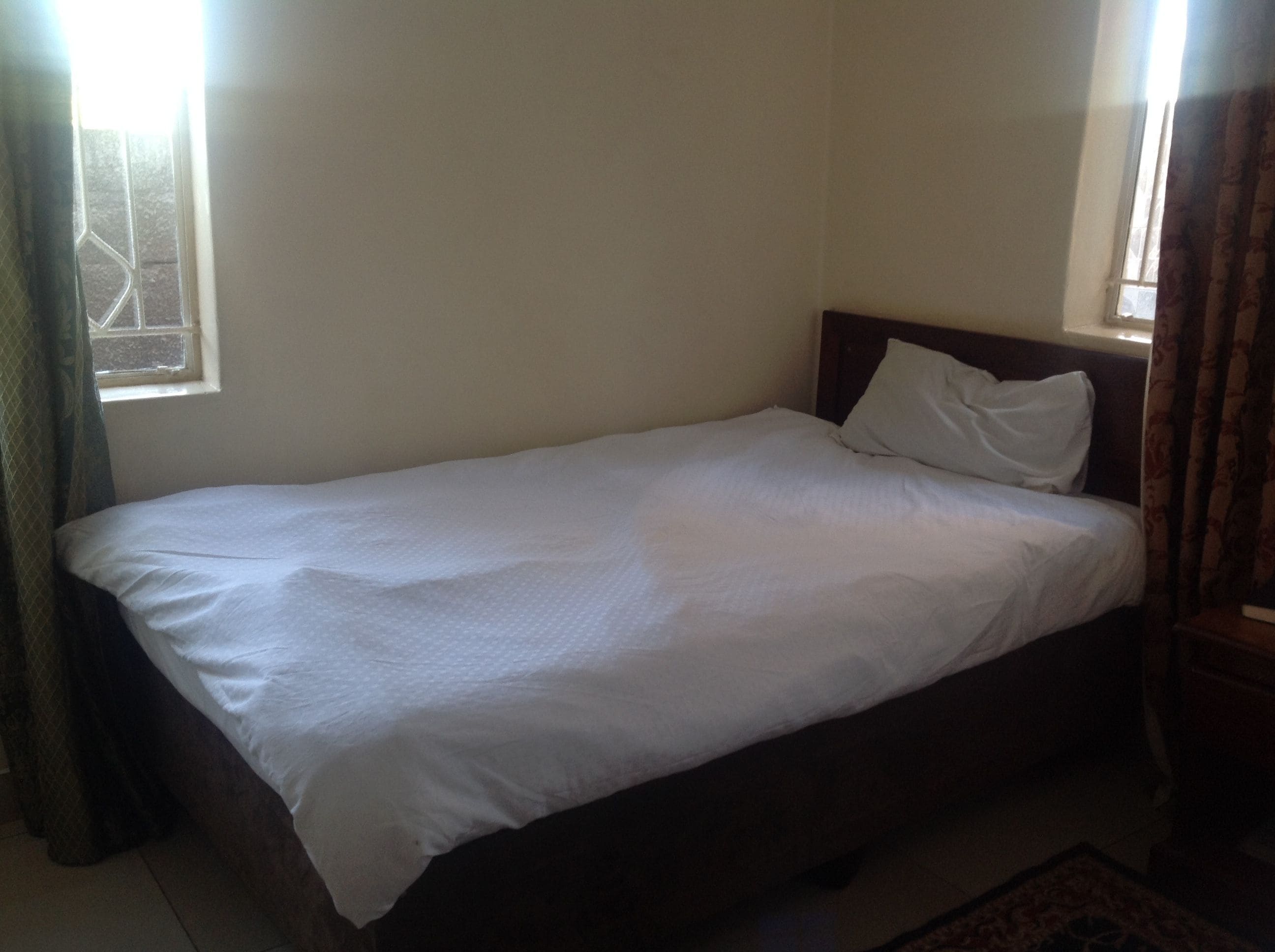 A single bed with white bedding in a room with plain walls and a window with curtains.