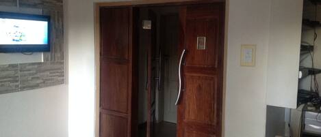 Una habitación con puerta de madera, un televisor fijado a la pared y una mesita con un documento encima.