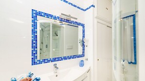A bathroom with a white sink, a mirror with a blue-tiled frame, a glass shower enclosure, and a towel rack. - Marino Suite Sorrento (Sorrento)