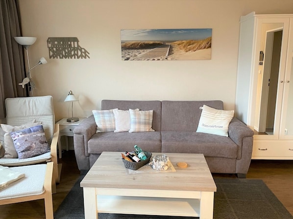 Ein Wohnzimmer mit grauem Sofa, zwei Kissen, einem Couchtisch mit Flaschen und einem Korb, einer Lampe und einem Gemälde einer Strandansicht.