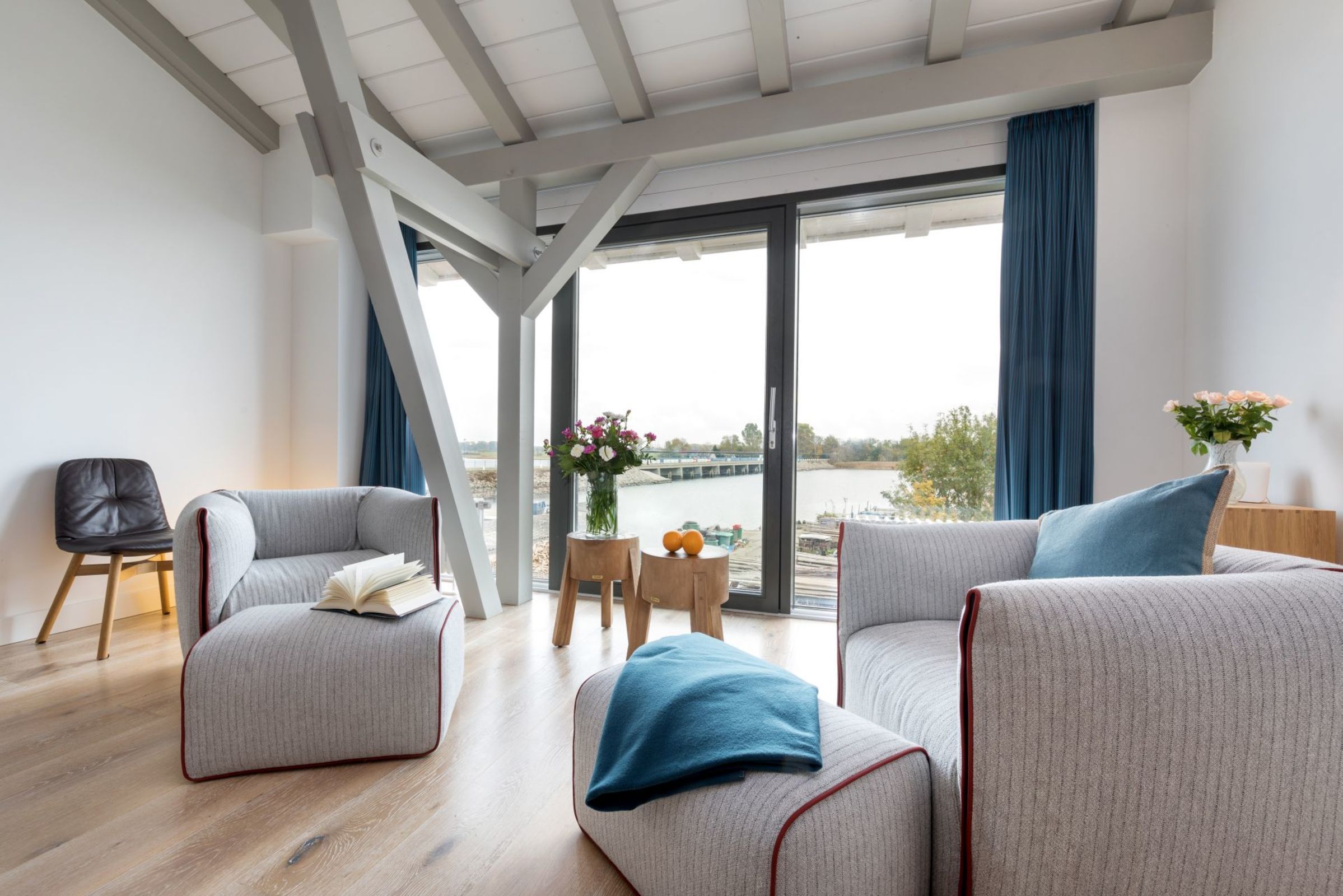 A modern living room with a sofa, ottoman, and chair, a small table with a vase, and a view of a river through large windows.