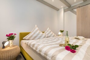 A neatly made bed with striped bedding, a bedside table with a vase of flowers, a bottle of champagne, and glasses. - AS1C - Old barn (AS) at c a l l s e n - appartements (Ummanz)