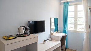 Habitación de hotel con televisor, escritorio con cafetera, espejo y ventana con cortinas.