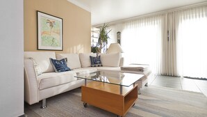 Ein modernes Wohnzimmer mit einem Glastisch, einem weißen Sofa mit blauen und weißen Kissen, einer großen Kübelpflanze und Blick ins Freie durch leichte Vorhänge.