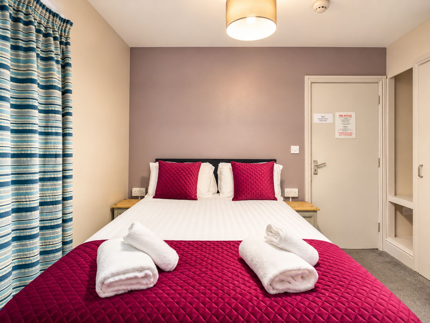 Habitación de hotel con cama, dos almohadas, colcha acolchada roja, dos toallas blancas, cortina a rayas y una puerta con aviso contra incendios.